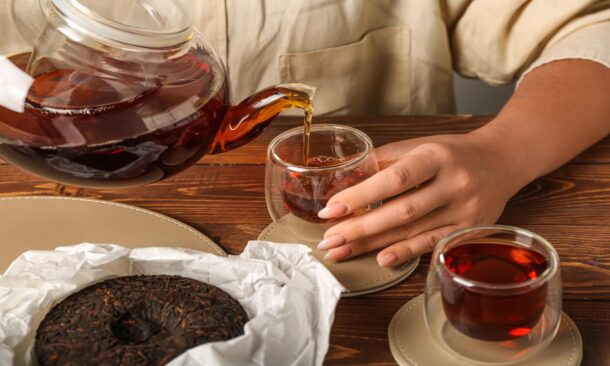 Dark tea being poured from a glass teapot into a small glass cup on a wooden table.