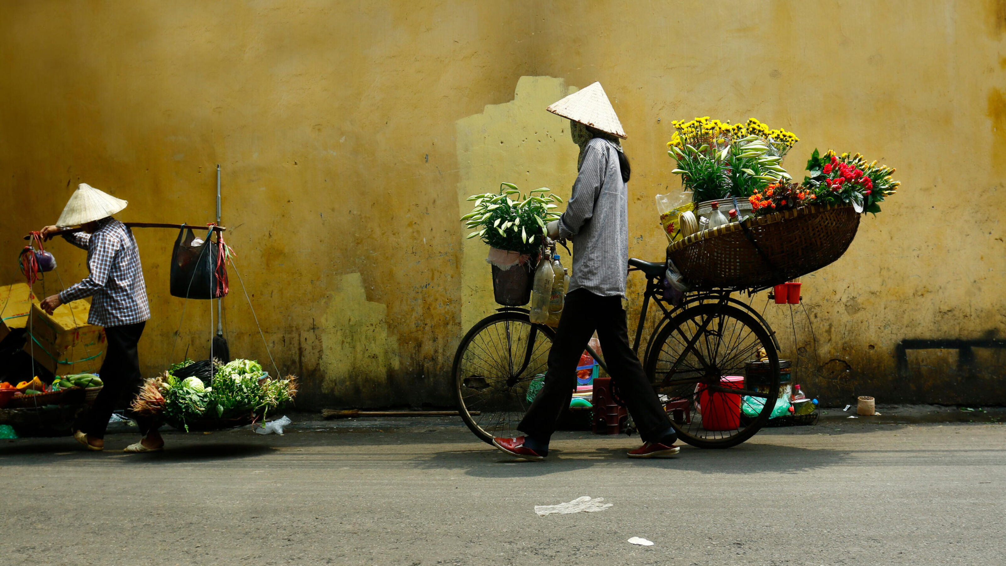 Two vendors in conical hats walking along a yellow wall, one with a bicycle and one with a shoulder pole.