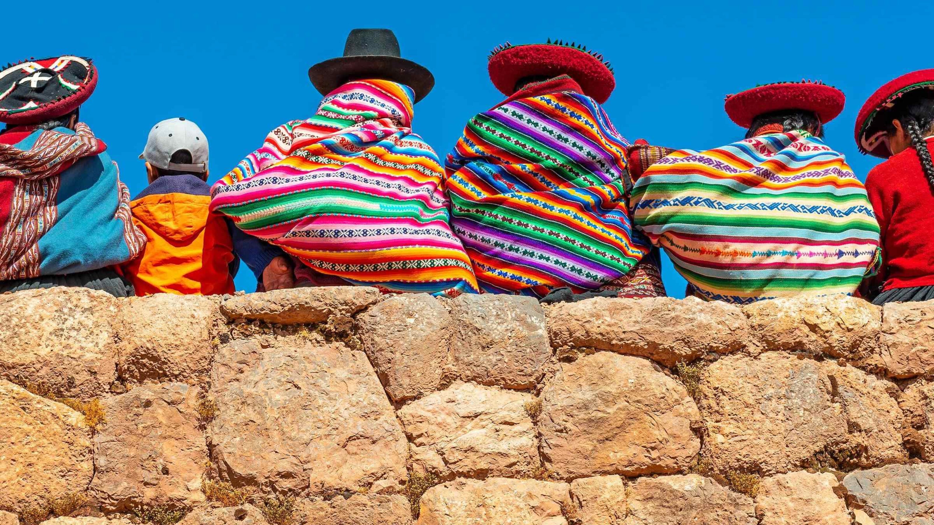 Panoramic photograph of Quechua indigenous women in traditional clothing with a boy sitting on an ancient Inca wall in Chinchero, Cusco Province, Peru.