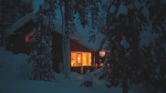 A dark wooden house with glowing yellow windows surrounded by snow-covered trees at dusk.