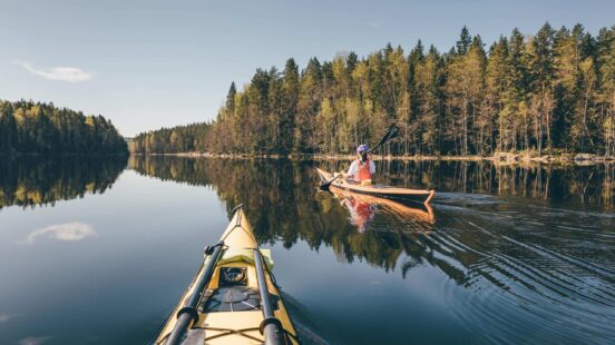 POV from a yellow kayak on a still lake with another person kayaking ahead near a forested shoreline.