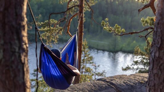 A person resting in a blue hammock tied between trees on a cliff overlooking a quiet lake.