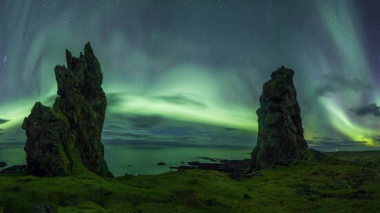 Green northern lights swirl over two jagged rock formations by the ocean under a starry sky.