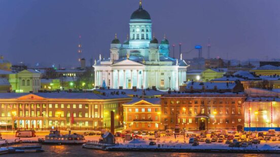 A white domed cathedral glows at night above a snowy city square with parked cars and harbor.
