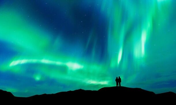 Silhouette of two people standing on a hill under a massive, glowing green and blue aurora.