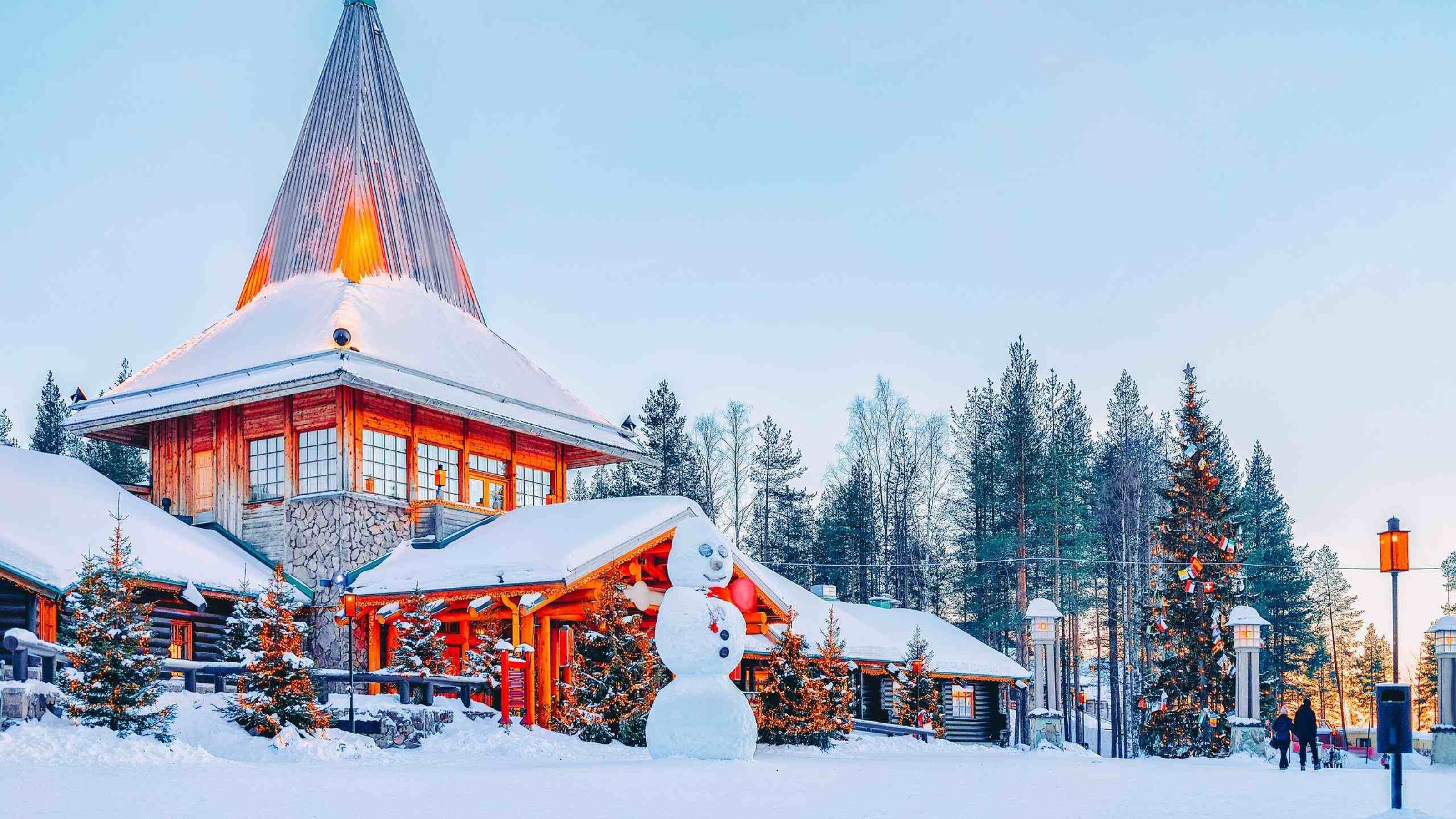A giant snowman in front of a lit wooden cabin with a tall steeple in a snowy forest.