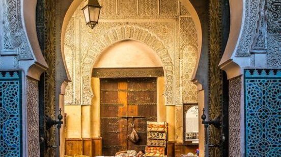 A highly detailed carved stone archway frames an entrance to a courtyard with a merchantx27;s stall.