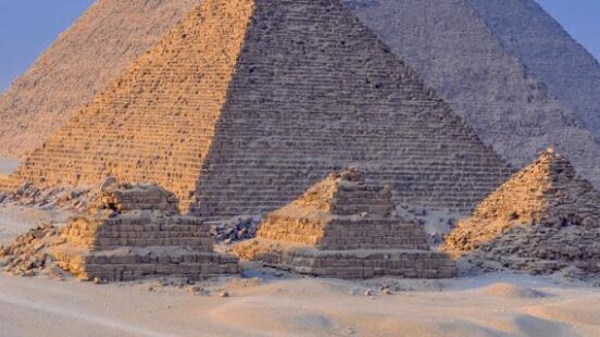 Three large ancient pyramids rise from the desert sand under a clear blue sky with camels in the distance.