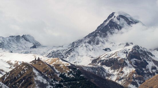 A high, snow-covered mountain peak rises above a small stone church on a hillside shrouded in clouds.