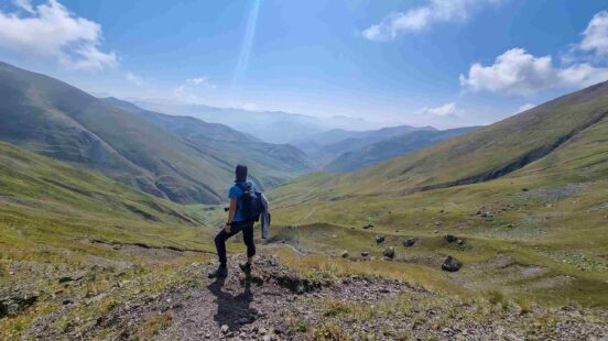 A hiker with a backpack stands on a rocky peak overlooking a wide mountain valley with rolling green slopes.
