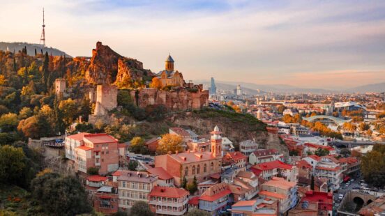 View of an old city with traditional architecture and a stone fortress on a hill against a sunset sky.