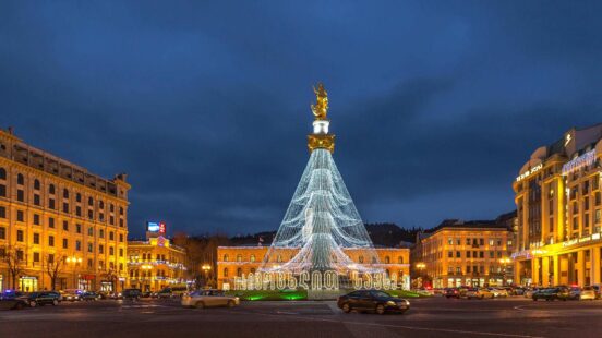 A city square at twilight with a tall monument covered in white lights and surrounding yellow-lit buildings.