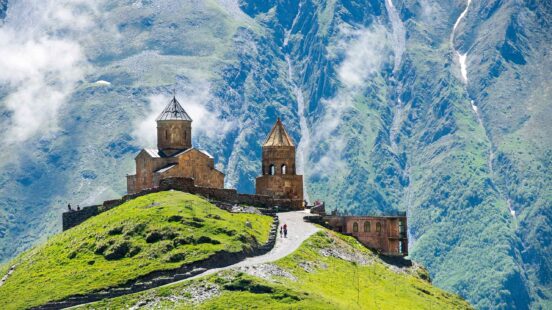 A stone church and tower on a green hill with large mountains in the background.