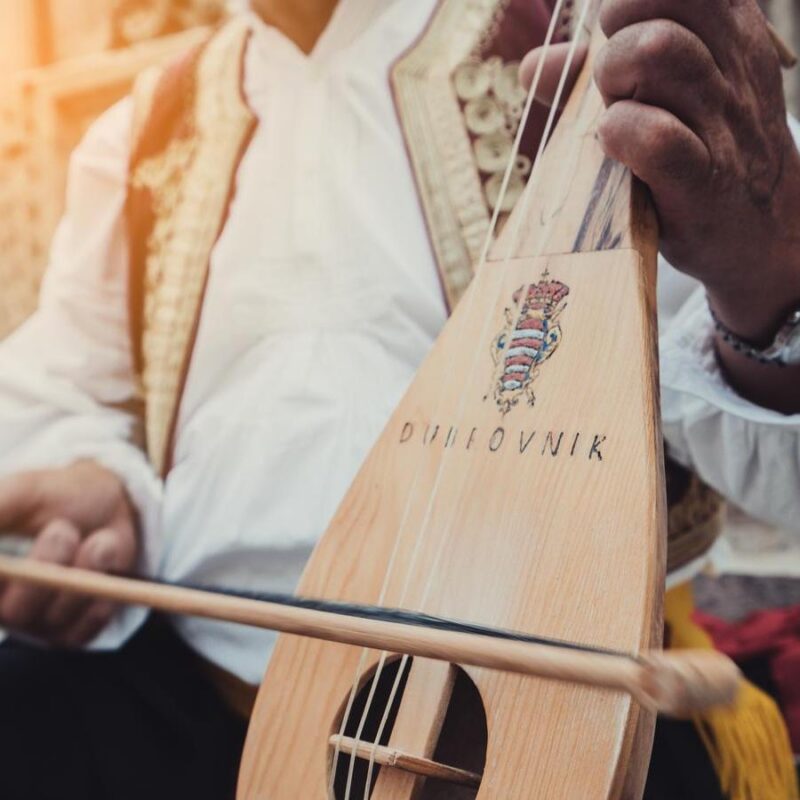 A busker plays traditional Croatian folk music with a 3-string instrument (lijerica) in the old city of Dubrovnik, Croatia. Man Plays Croatian Musical Instrument in Dubrovnik art, artist, band, busker, city, croatia, croatian, culture, dubrovnik, dubrovnik old town, dubrovnik street, entertainment, event, festival, folk, fun, instrument, jazz, lifestyle, lijerica, live, male, men, music, music street performers, musical, musician, old, outdoor, people, perform, performance, performer, person, play, player, playing, public, sound, street, street busker, street style, string, tourism, town, tradition, traditional, travel, urban