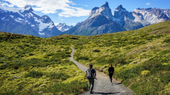 Two people hiking on a trail in a vast mountain landscape with jagged peaks and green vegetation.