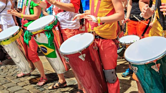 Close-up of people playing tall drums on a cobblestone street, featuring a small Brazilian flag.