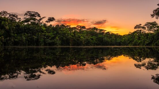 A sunset sky with orange clouds reflecting on a still lake surrounded by rainforest.