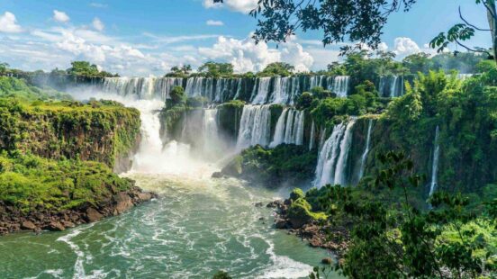 Wide view of many waterfalls flowing over verdant cliffs in a jungle setting.