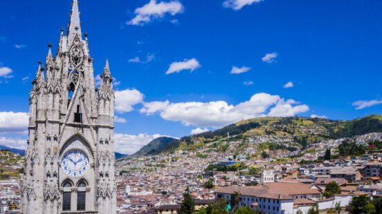 A tall gothic stone cathedral tower with a clock overlooking a city and mountains.