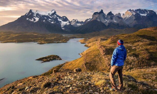 Hiker in a blue jacket looking out over a mountain lake and rugged peaks at sunset.