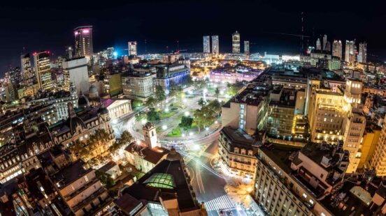 Nighttime cityscape featuring a glowing central square, street lights, and various architectural styles.