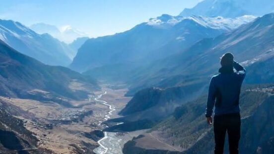 Hiker overlooking a deep mountain valley with a winding river and distant snowy peaks.