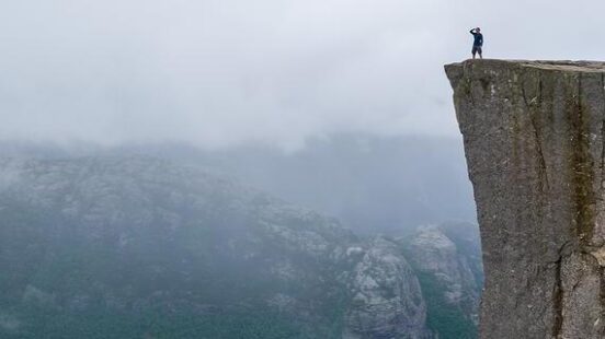 Hiker standing on the edge of a flat rock cliff overlooking a bright blue fjord and misty mountains.