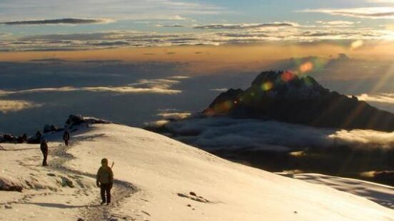 People hiking on a snowy mountain ridge above the clouds during a golden sunset.