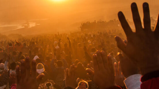 Many people raise their hands toward a glowing sun over a wide, misty landscape at dawn or dusk.