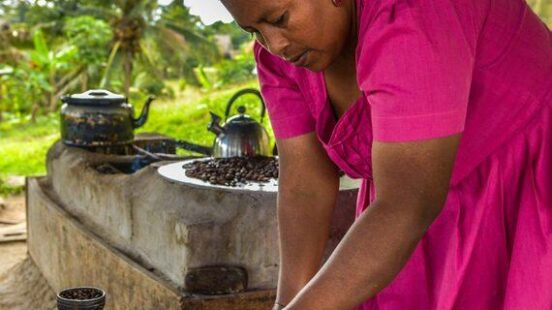 A woman in a bright pink dress stands under a thatched roof, processing roasted cacao beans on a round wooden table. Part of luxury Belize tours.