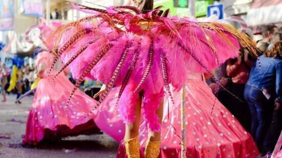 A dancer in a large, pink feathered costume and gold boots performs on a confetti-strewn street.