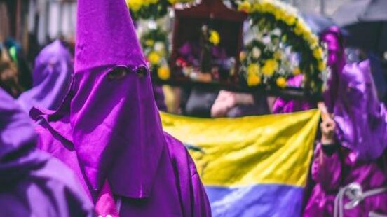 A participant in a pointed purple hood and robe stands during a street procession with a flag in the background.