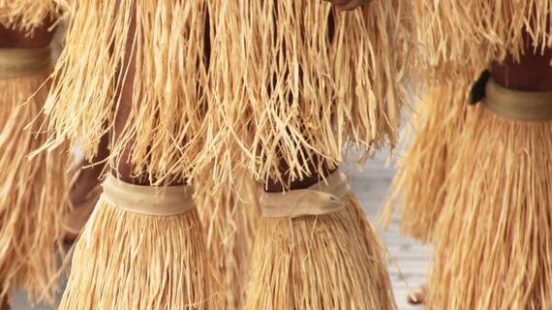 Close-up of dancers wearing skirts and leg bands made of long, tan natural fibers.