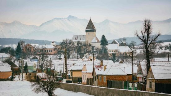 A view of a fortified church with a tall spire and surrounding village structures covered in snow, set against a backdrop of snowy mountains.