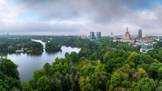 Panoramic view of a city with a large lake and dense green trees in the foreground and tall buildings in the background.