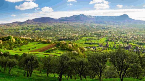 Wide landscape of green rolling hills, a valley with small settlements, and a rugged mountain range under a blue sky.