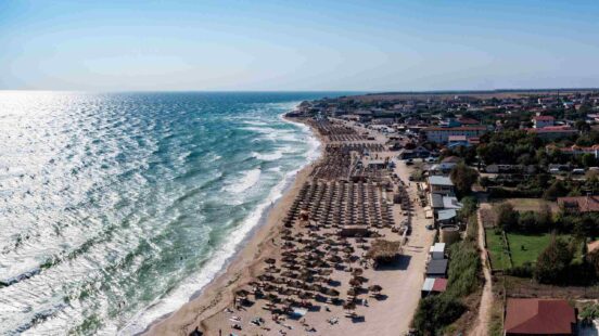 Aerial view of a long, crowded beach with umbrellas and sunbeds, next to the wavy ocean and a coastal town.
