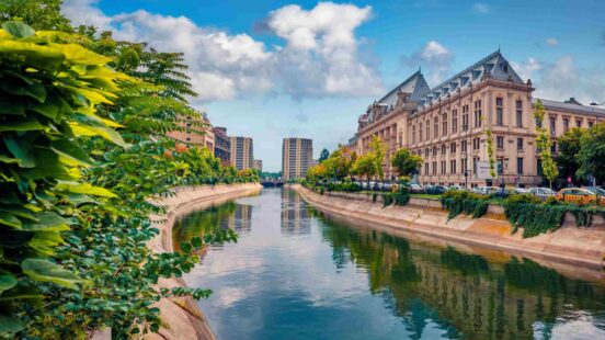 A river flowing through a city, flanked by modern and historic buildings, with lush green trees lining the banks.