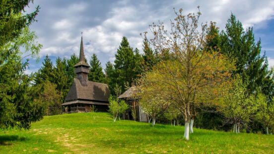 Traditional wooden church with a tall spire on a grassy hill, surrounded by coniferous trees under a partly cloudy sky.