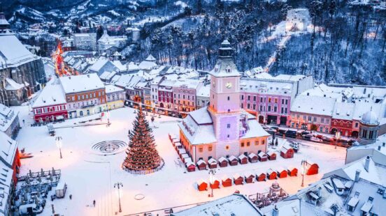 Aerial view of a snowy town square at dusk with a clock tower, a lit Christmas tree, and surrounding historic buildings.