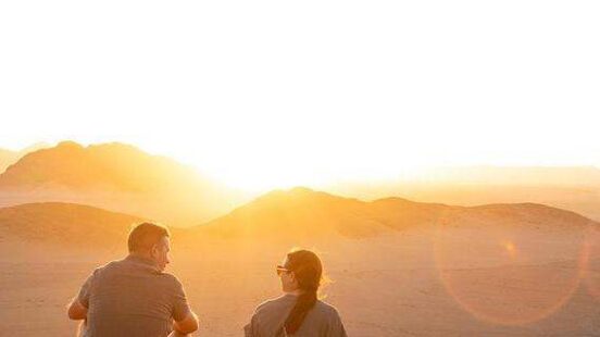 Silhouette of two people sitting on a rock watching a bright sunset over desert mountains.