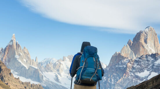 Rear view of a hiker with a backpack looking at tall, rocky mountain peaks under a blue sky.