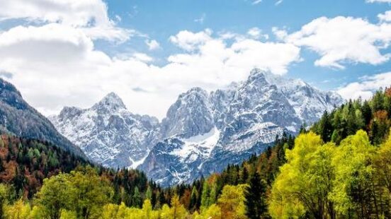 Snow-capped mountain range reflecting in a clear lake with green trees and a blue cloudy sky.