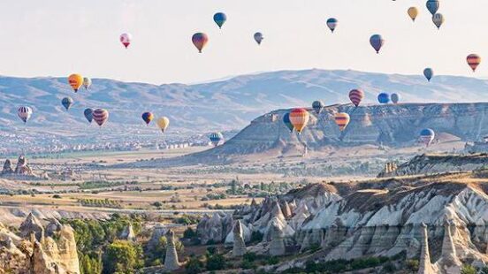 Many colorful hot air balloons fly over a rocky valley with unique geological formations under a clear sky.