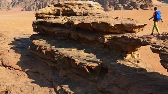 A person with a blue backpack walks on a rock formation in a vast desert landscape with large sandstone mountains.