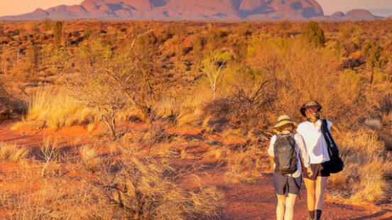 Two people walk on a red trail toward large rounded rock formations under a soft pink and blue sunset sky.