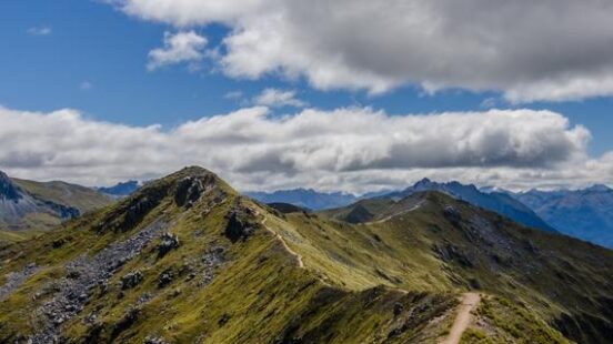A narrow dirt path winds along a steep green mountain ridge under a blue sky with white clouds.