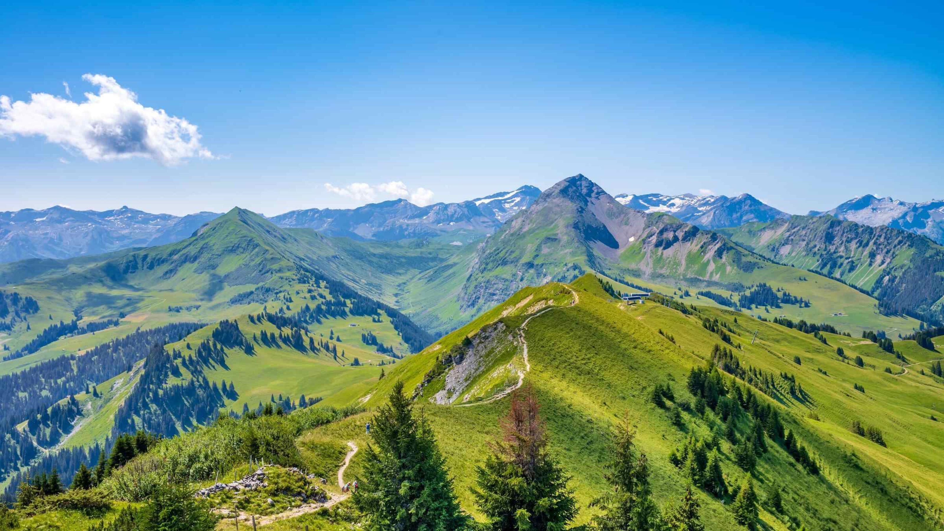 Rolling green hills and jagged mountains under a bright blue sky with a small white cloud.
