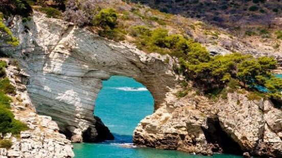 Gargano national park in Puglia, Italy, with a dramatic rock archway eroded out of the coastline through which bright blue sea is visible