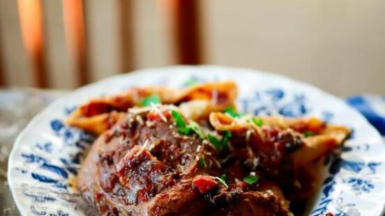 Braised meat and pasta garnished with cheese and herbs on a decorative plate next to a glass of wine.